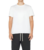 A person dressed in a Frame Men's Logo Tee by Frame and black drawstring pants is standing against a plain background.