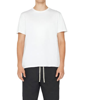 A person dressed in a Frame Men's Logo Tee by Frame and black drawstring pants is standing against a plain background.