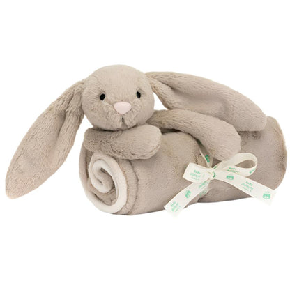 A Jellycat Bashful Beige Bunny Blankie, crafted with recycled fibers, features a stuffed beige bunny with long ears resting on a rolled-up matching beige blanket secured with a ribbon that has green text.