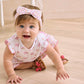 A baby girl with blue eyes wears the Kissy Kissy Baby Girls’ Headband—a soft, stretchy bow accessory—while crawling on a light wood floor in a white outfit with pink bows and brown sandals.