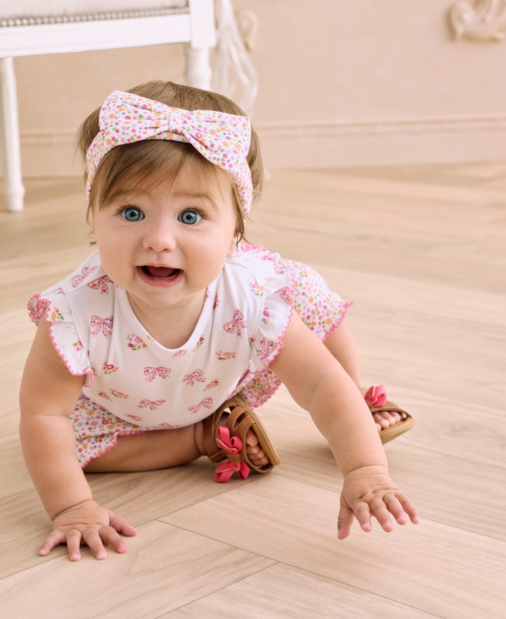 A baby girl with blue eyes wears the Kissy Kissy Baby Girls’ Headband—a soft, stretchy bow accessory—while crawling on a light wood floor in a white outfit with pink bows and brown sandals.