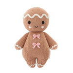 Baby Gingerbread - Candy Swirl by Cuddle + Kind is a knitted gingerbread doll handcrafted from Peruvian cotton, featuring a smiling face, white icing details, and two charming pink bow decorations on its chest—an irresistibly soft companion for babies, pictured against a white background.