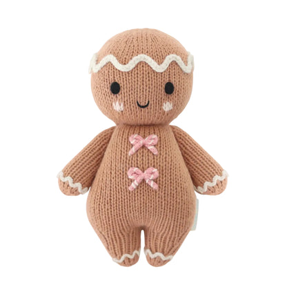 Baby Gingerbread - Candy Swirl by Cuddle + Kind is a knitted gingerbread doll handcrafted from Peruvian cotton, featuring a smiling face, white icing details, and two charming pink bow decorations on its chest—an irresistibly soft companion for babies, pictured against a white background.
