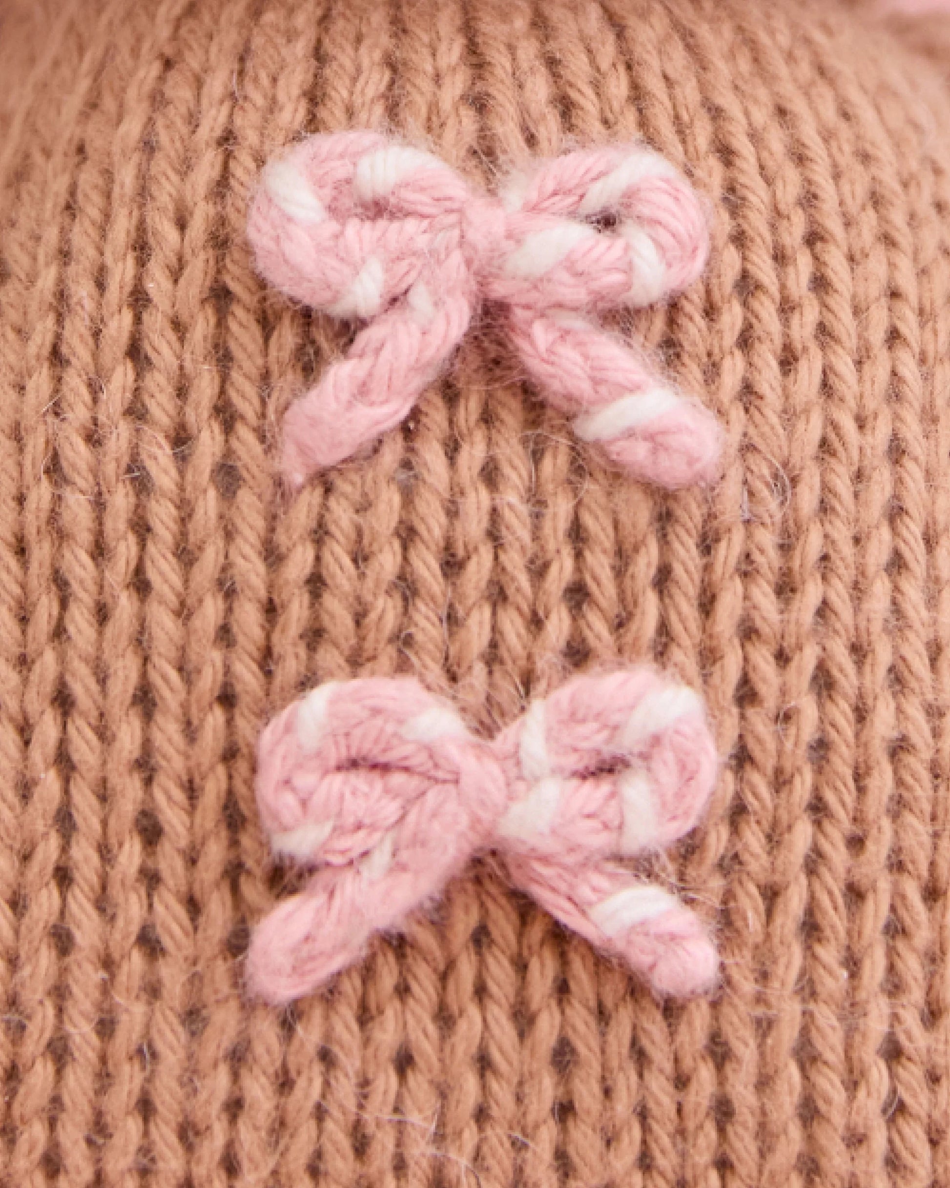 Close-up of the Baby Gingerbread - Candy Swirl by Cuddle + Kind, showcasing a knitted brown fabric with two small pink and white yarn bows, meticulously handcrafted from heirloom-quality Peruvian cotton for a soft and cherished baby companion.