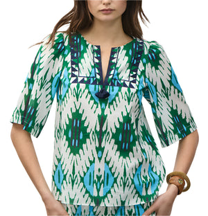 A woman wears the Figue Bodhi Top, a patterned green, blue, and white cotton voile blouse with elbow-length sleeves and delicate embroidery highlighting the geometric V-neck design.