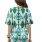 A woman is seen from behind wearing the Figue Bodhi Top, a short-sleeved cotton voile blouse with a green, blue, and white geometric pattern. She accessorizes with a bracelet on her right wrist.