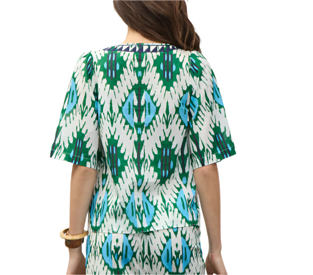 A woman is seen from behind wearing the Figue Bodhi Top, a short-sleeved cotton voile blouse with a green, blue, and white geometric pattern. She accessorizes with a bracelet on her right wrist.