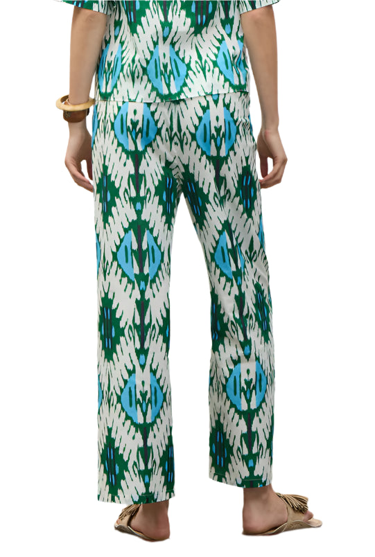 A person stands with their back to the camera, wearing Figue's Justine Pant featuring a blue and green geometric print, side pockets, and a drawstring waist, paired with the matching top, tan sandals, and a chunky bracelet.