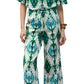 A person wearing the Figue Justine Pant, featuring a blue, green, and white pattern with a drawstring waist and side pockets, paired with sandals and a bracelet, stands against a white background.