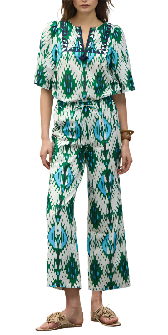 A person wearing the Figue Justine Pant, featuring a blue, green, and white pattern with a drawstring waist and side pockets, paired with sandals and a bracelet, stands against a white background.