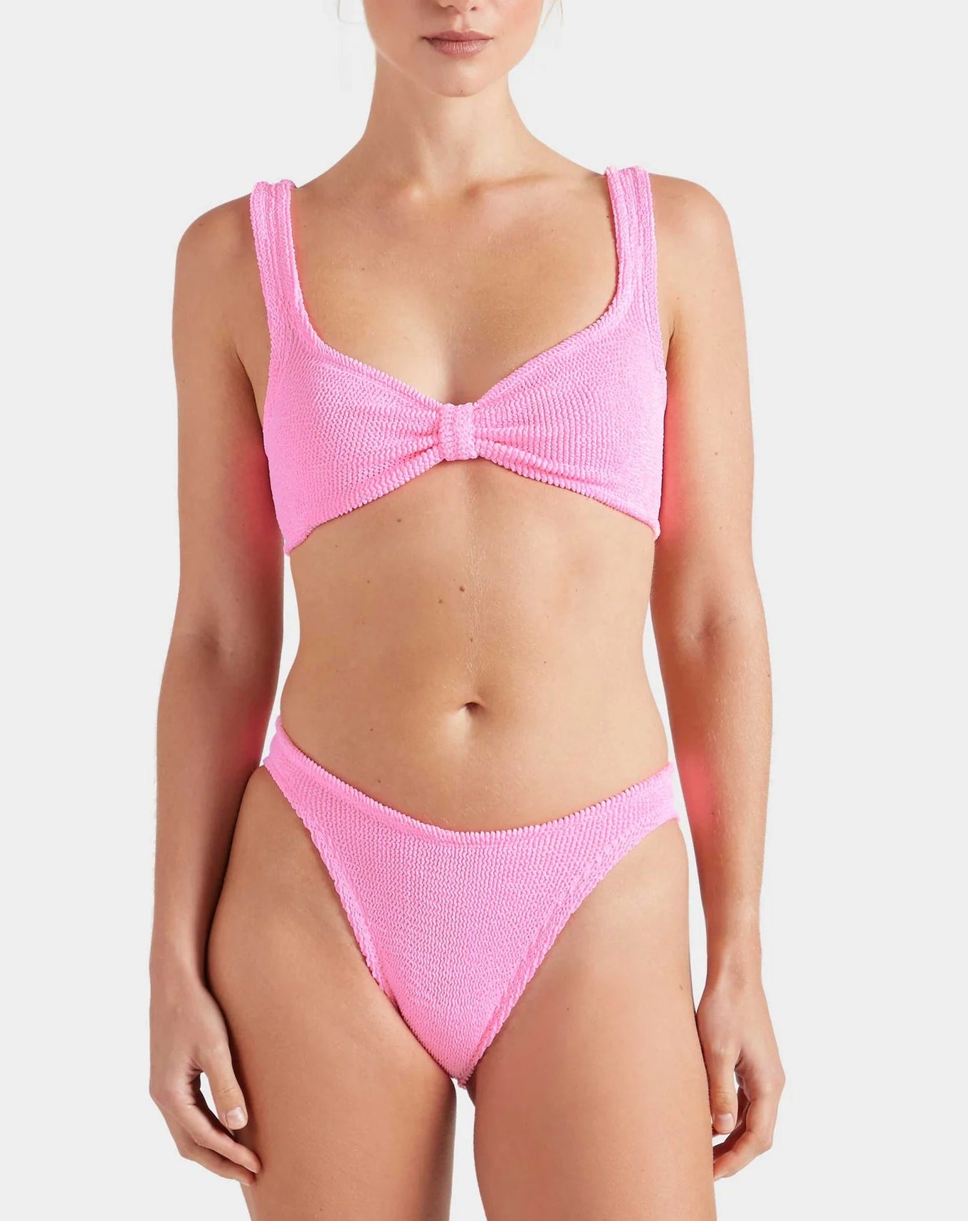 Model wears the Bonnie Bikini by Hunza G—a textured light pink retro set with a knotted, supportive top and high-waist briefs—against a plain light background.