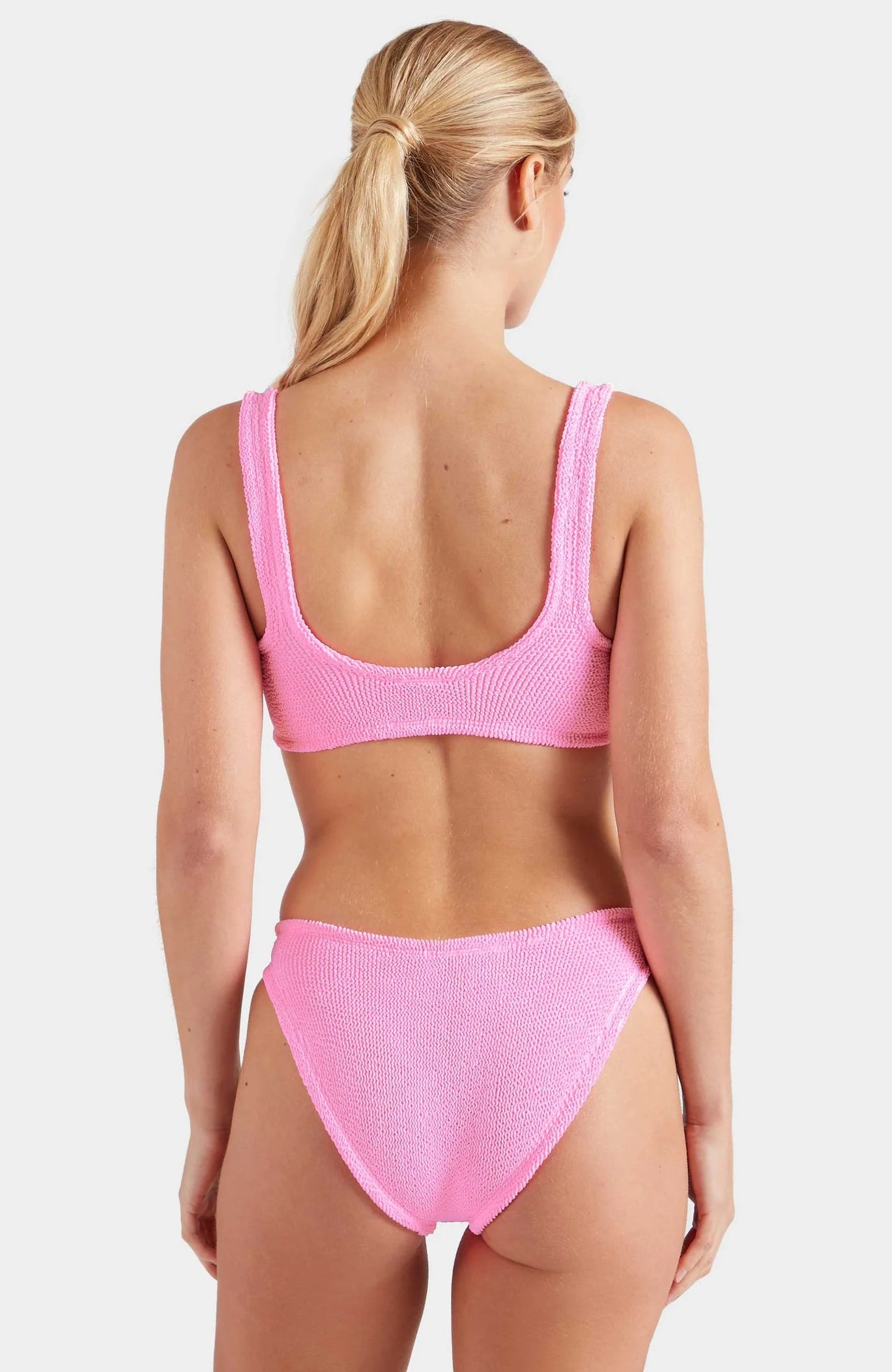 A woman with blonde hair in a ponytail is seen from behind wearing the textured retro Hunza G Bonnie Bikini with high waist briefs, set against a plain white background.