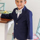 A young boy in The Beaufort Bonnet Company's Boys' Booker Blazer, pastel bow tie, and white shorts stands indoors beside a table, smiling at the camera in preppy perfection.