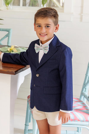 A young boy in The Beaufort Bonnet Company's Boys' Booker Blazer, pastel bow tie, and white shorts stands indoors beside a table, smiling at the camera in preppy perfection.