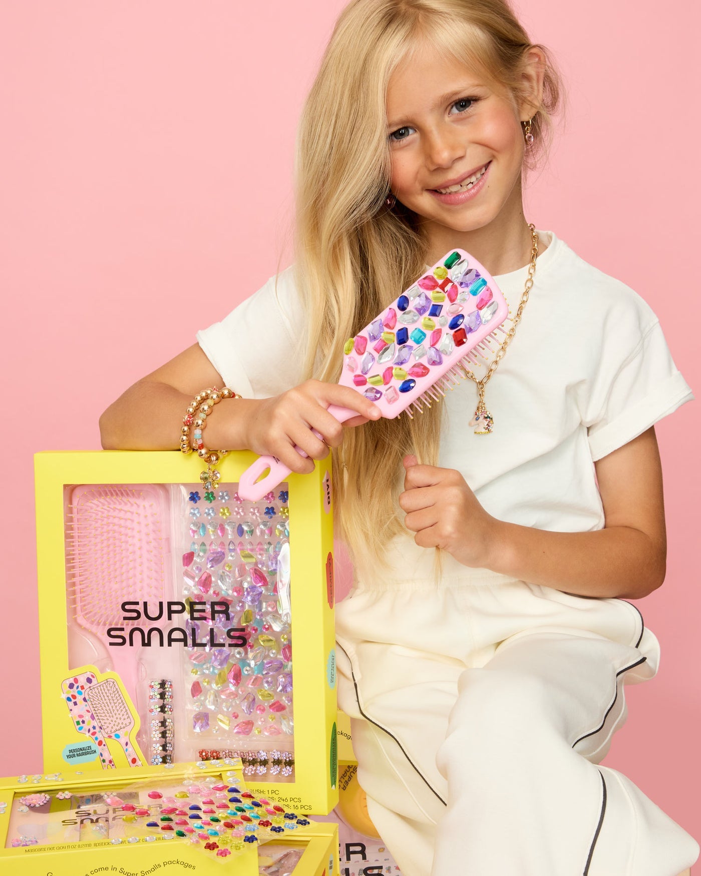 A smiling girl in a white outfit holds a colorful kids hairbrush, sitting next to a boxed Super Smalls Style and Shine Gemify Hairbrush Kit, against a pink background.