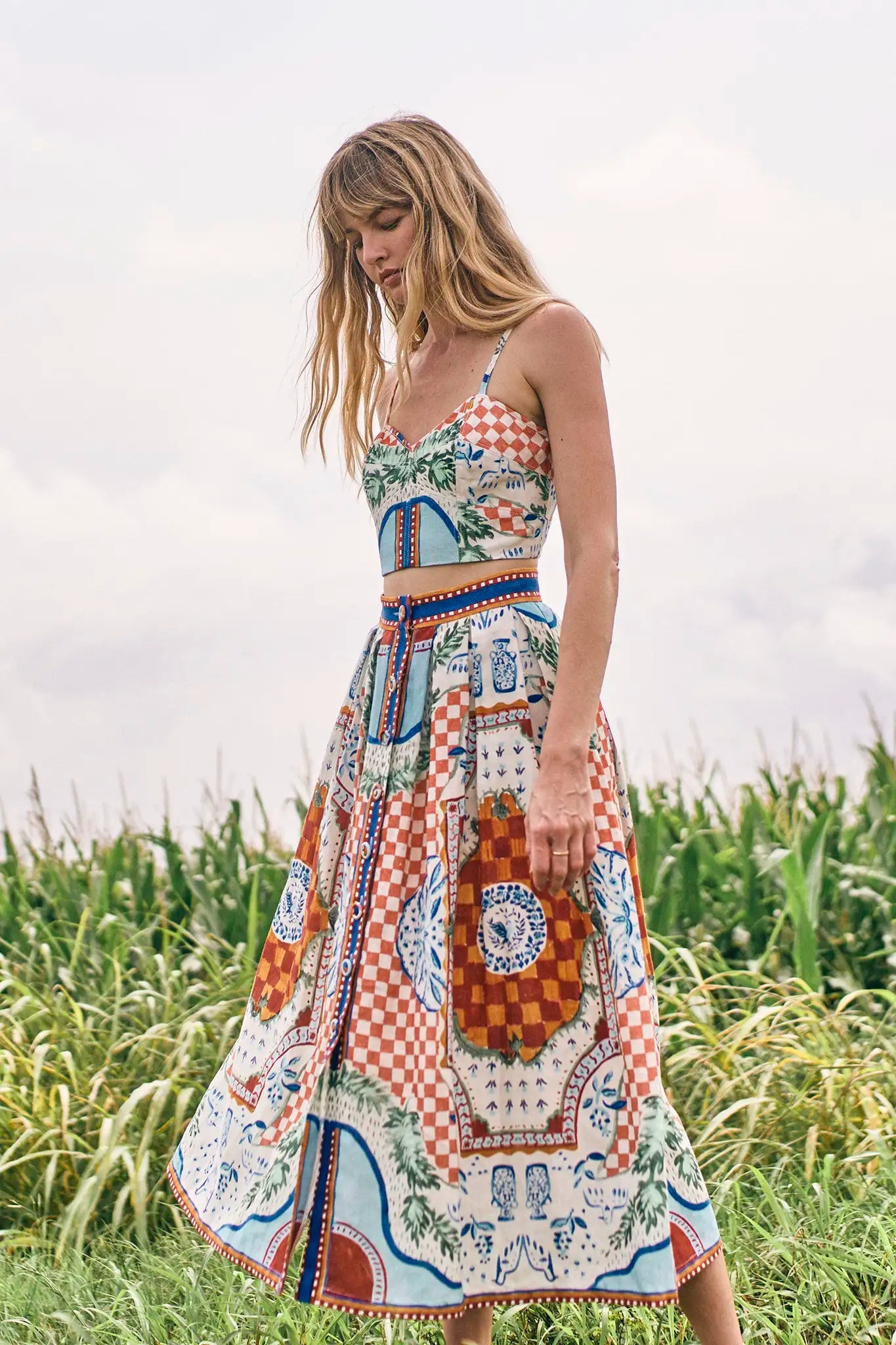 A woman stands outdoors before tall green corn plants on a cloudy day, wearing the Eva Skirt by Hunter Bell NYC in a colorful sundance tile print, crafted from a comfy linen-rayon blend.
