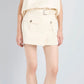 A person stands against a light background wearing the Proenza Schouler Bruni Short—a sleeveless beige top paired with matching high-rise shorts featuring front pockets and a belted waist—and black open-toe sandals.