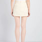A person stands with their back to the camera, wearing a white sleeveless top, the Bruni Short by Proenza Schouler in beige with belt loops and a wrap-front design, paired with black sandals against a plain light background.