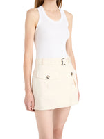 A woman wearing a white sleeveless tank top and the Bruni Short by Proenza Schouler—cream high-rise shorts featuring a belted waist, front flap pockets, and button details—standing against a plain background.