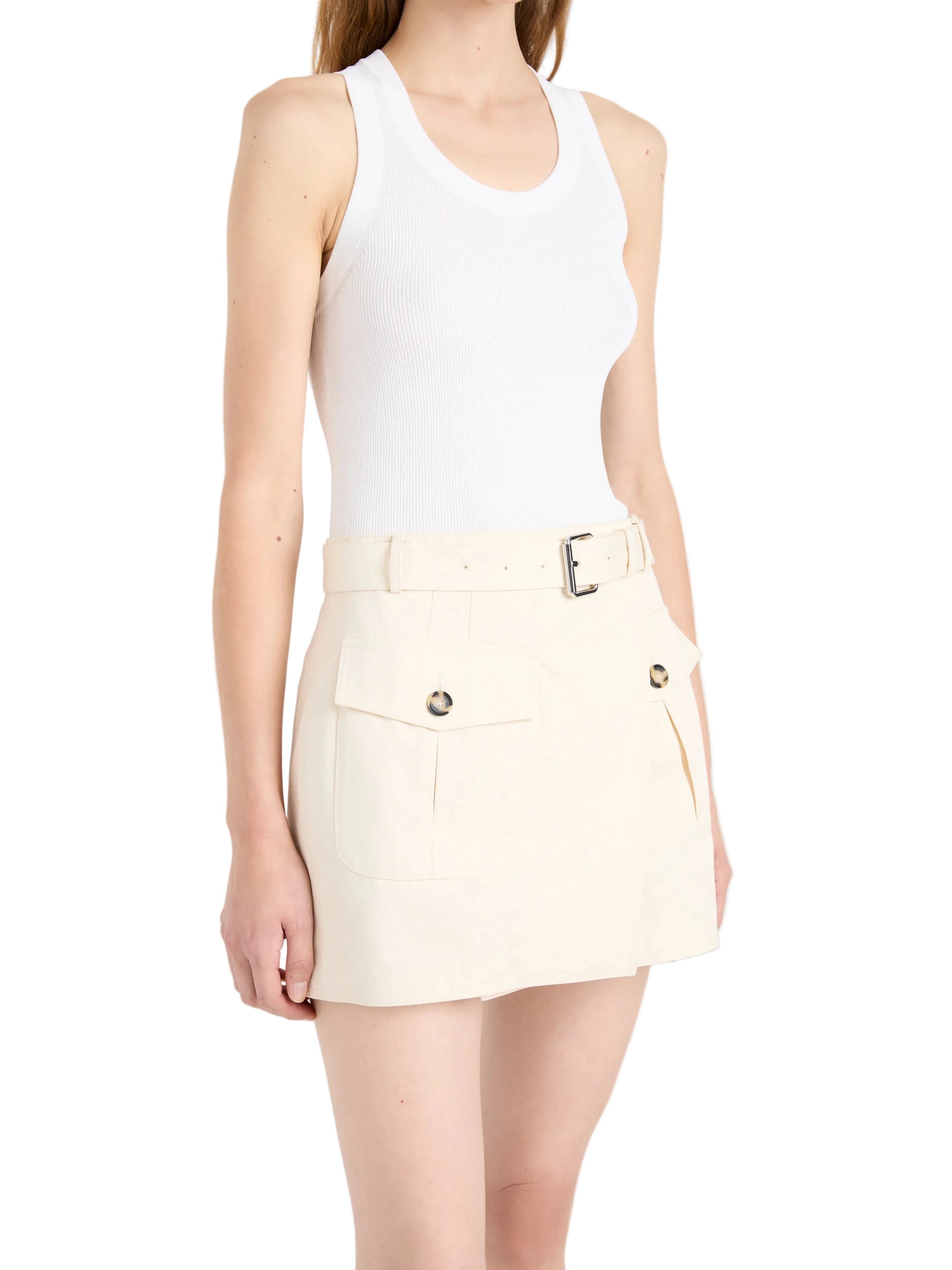 A woman wearing a white sleeveless tank top and the Bruni Short by Proenza Schouler—cream high-rise shorts featuring a belted waist, front flap pockets, and button details—standing against a plain background.