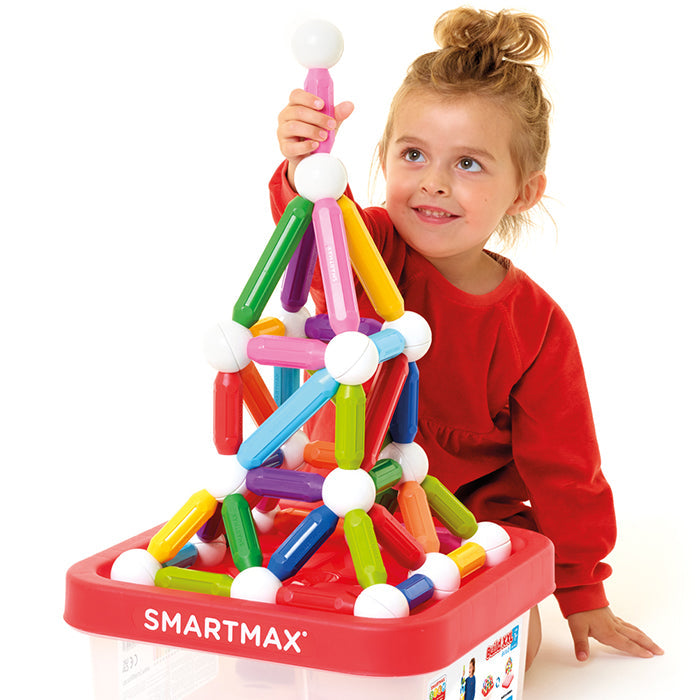 A young child in a red shirt sits beside a bin labeled "SMARTMAX," building colorful structures with SmartMax Build XXL magnetic rods and balls from Smart Games.