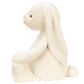 A Jellycat Bashful Cream Bunny, Giant with long ears sits upright, facing left against a plain background. Crafted from soft polyester, this charming stuffed bunny by Jellycat adds a cozy and delightful touch.