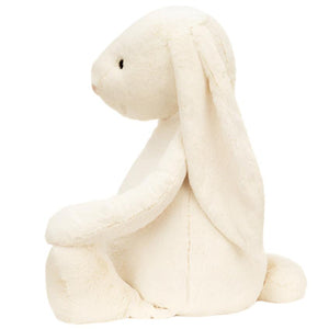 A Jellycat Bashful Cream Bunny, Giant with long ears sits upright, facing left against a plain background. Crafted from soft polyester, this charming stuffed bunny by Jellycat adds a cozy and delightful touch.