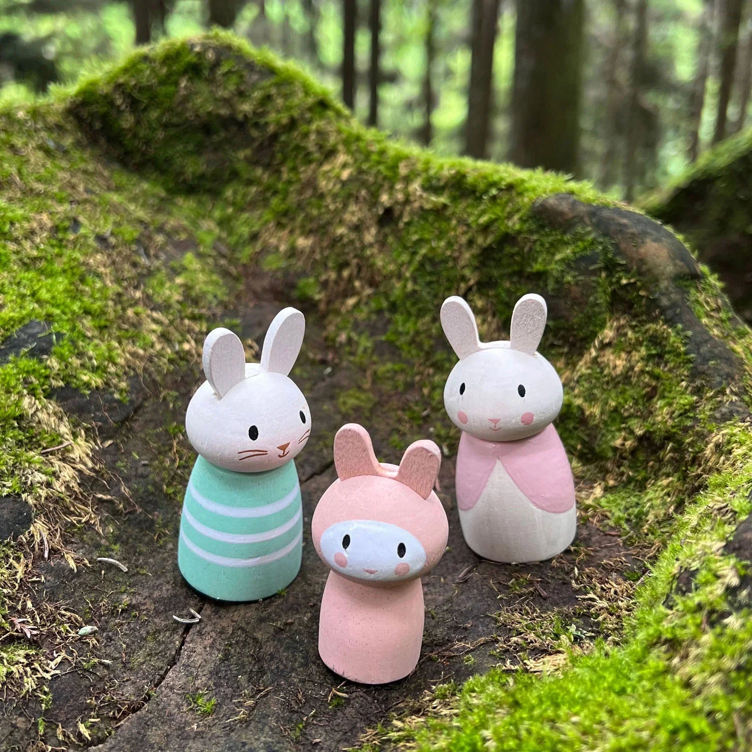 The Tender Leaf Bunny Tales set from Tender Leaf Toys features three wooden rabbit figurines, representing a delightful bunny family. They stand on a mossy tree stump in a forest setting, with their colorful outfits adding whimsy to this dollhouse scene.