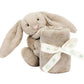 A Jellycat Bashful Beige Bunny Soother, crafted from recycled fibers, holds a rolled-up beige baby blanket tied with a cream-colored ribbon.