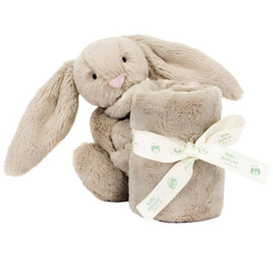 A Jellycat Bashful Beige Bunny Soother, crafted from recycled fibers, holds a rolled-up beige baby blanket tied with a cream-colored ribbon.