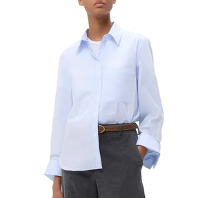 Wearing the TWP Boyfriend Shirt in light blue Italian cotton over a white top, paired with grey pants and a brown belt, this person stands with one hand in their pocket.