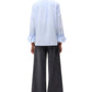 Person standing with back to camera, wearing the TWP Boyfriend Shirt in light blue Italian cotton with rolled-up sleeves, paired with wide-leg dark gray pants.