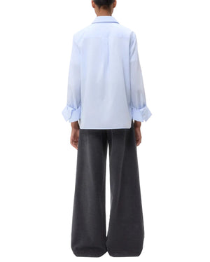 Person standing with back to camera, wearing the TWP Boyfriend Shirt in light blue Italian cotton with rolled-up sleeves, paired with wide-leg dark gray pants.