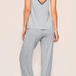 A woman stands barefoot facing away, wearing Eberjey’s Frida Cami & Pant PJ Set in light gray TENCEL Modal with black trim, featuring a soft camisole and matching loose pajama pants against a neutral background.