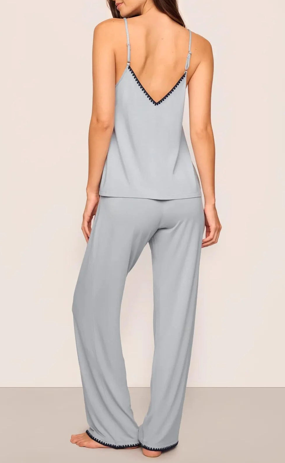 A woman stands barefoot facing away, wearing Eberjey’s Frida Cami & Pant PJ Set in light gray TENCEL Modal with black trim, featuring a soft camisole and matching loose pajama pants against a neutral background.