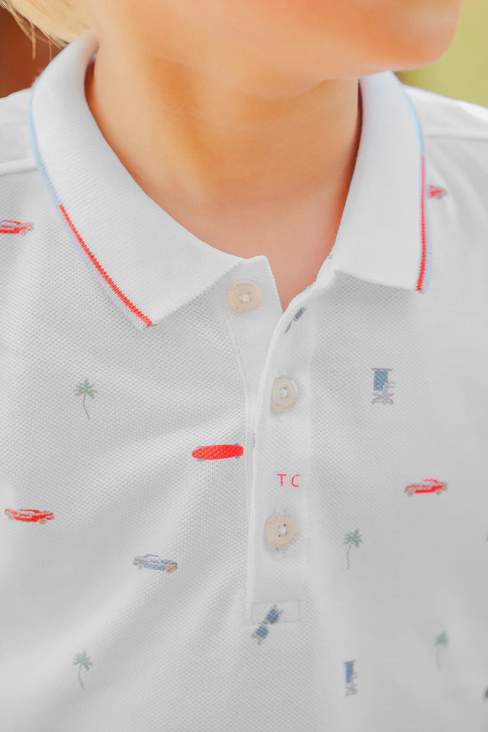 A close-up of Tartine et Chocolat's "Baby Boys' and Boys' Spicy Orange Polo" displays Miami-inspired motifs with palm trees, cars, and boats. The shirt has a red and light blue-lined collar and beige buttons.
