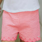 A child wearing Tartine et Chocolat's Baby Girls' and Girls' Coral Seersucker Shorts and a white shirt stands indoors, showcasing charm with every subtle pattern.