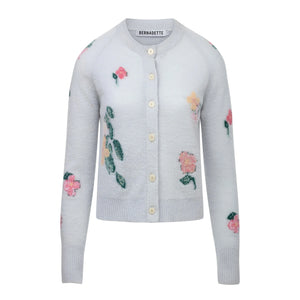 The Clemence Clematis Intarsia Cardigan by Bernadette features a soft wool knit in light blue-grey with a round neck, front buttons, and pink floral patterns, shown against a white background.