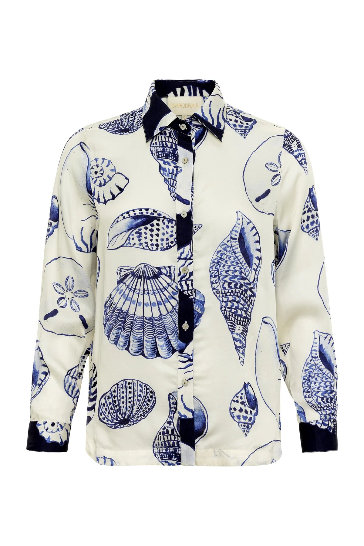 The Carmen Shirt by Carolina K is a long-sleeve button-up with a white base and blue seashell print, designed with South African artist Felicity Buchanan. It features a contrasting dark collar, placket, and cuffs.