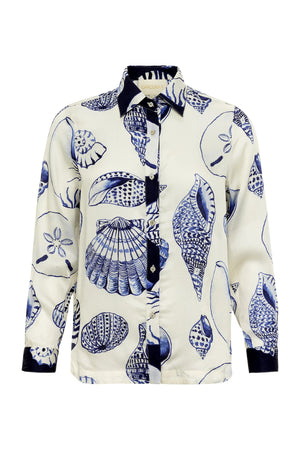 The Carmen Shirt by Carolina K is a long-sleeve button-up with a white base and blue seashell print, designed with South African artist Felicity Buchanan. It features a contrasting dark collar, placket, and cuffs.