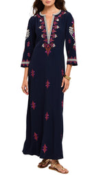 A woman wearing the Cassandra Dress by Figue—a navy blue long dress with pink and white embroidery, three-quarter sleeves, a V-neckline—paired with gold sandals.