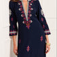 A woman wears the Cassandra Dress by Figue, featuring a navy blue color with pink and white embroidery, three-quarter sleeves, and a V-neckline with decorative trim.