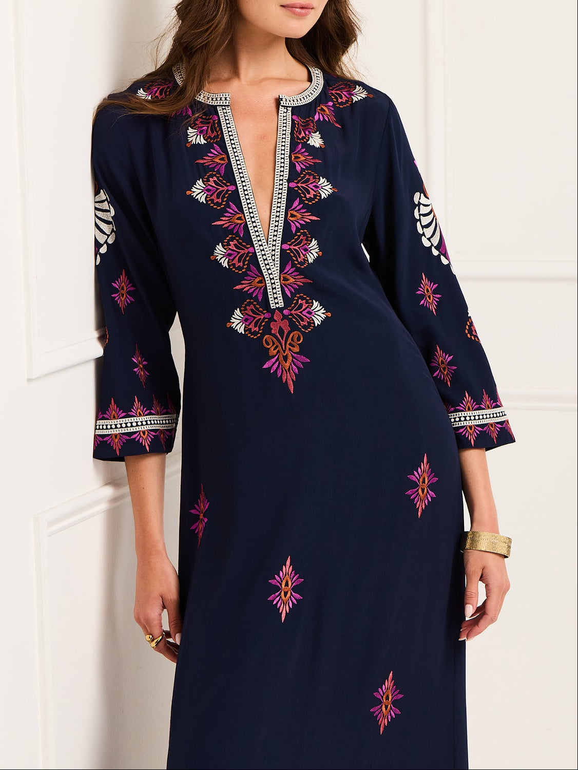 A woman wears the Cassandra Dress by Figue, featuring a navy blue color with pink and white embroidery, three-quarter sleeves, and a V-neckline with decorative trim.