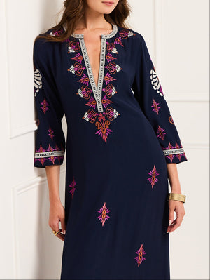 A woman wears the Cassandra Dress by Figue, featuring a navy blue color with pink and white embroidery, three-quarter sleeves, and a V-neckline with decorative trim.