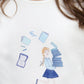A junior girl wears the Tartine et Chocolat Girls' Long-Sleeved Graphic T-Shirt in ecru, featuring a Parisian-inspired illustration of a girl with books and playful sketches of glasses, books, and a notebook.