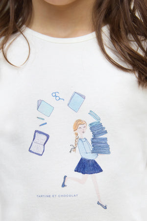 A junior girl wears the Tartine et Chocolat Girls' Long-Sleeved Graphic T-Shirt in ecru, featuring a Parisian-inspired illustration of a girl with books and playful sketches of glasses, books, and a notebook.