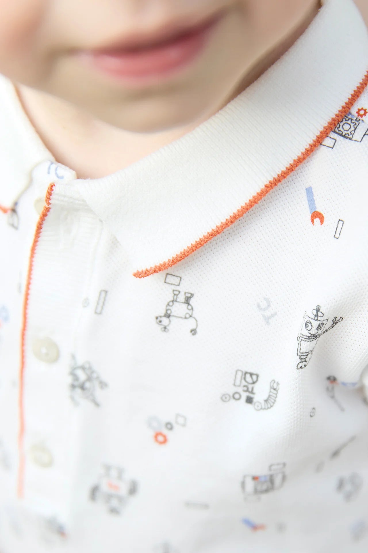 Close-up of a child in a Tartine et Chocolat Baby Boys' and Boys' Robots Illustration Long Sleeved Polo, showing the robot pattern, orange collar trim, and part of the child's face.