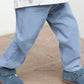 A young boy from the Tartine et Chocolat Baby Boys' and Boys' Trouser Pants collection stands on a stone surface, wearing blue-grey pants, a patterned white shirt, and blue shoes.