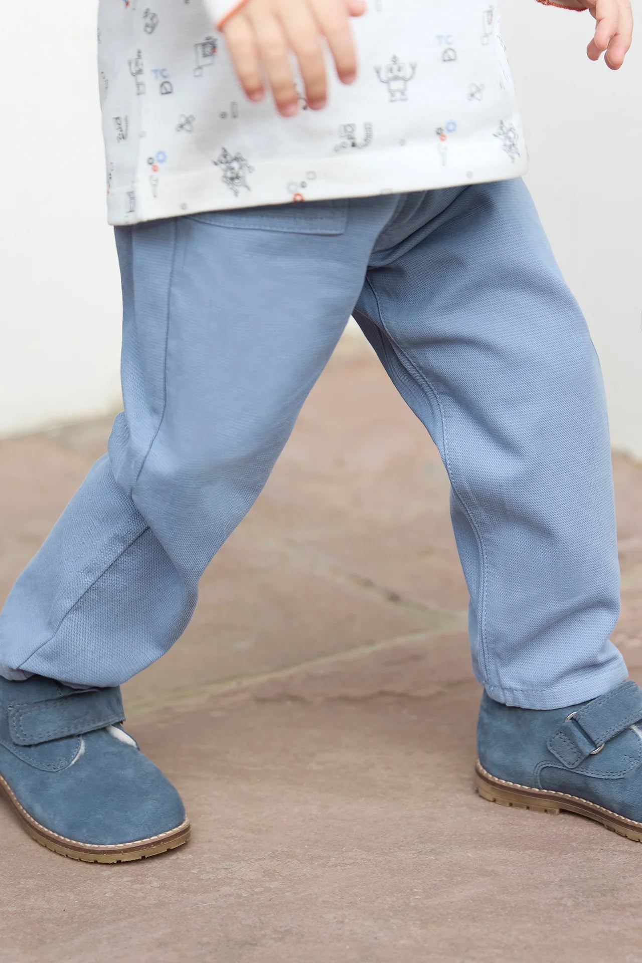 A young boy from the Tartine et Chocolat Baby Boys' and Boys' Trouser Pants collection stands on a stone surface, wearing blue-grey pants, a patterned white shirt, and blue shoes.