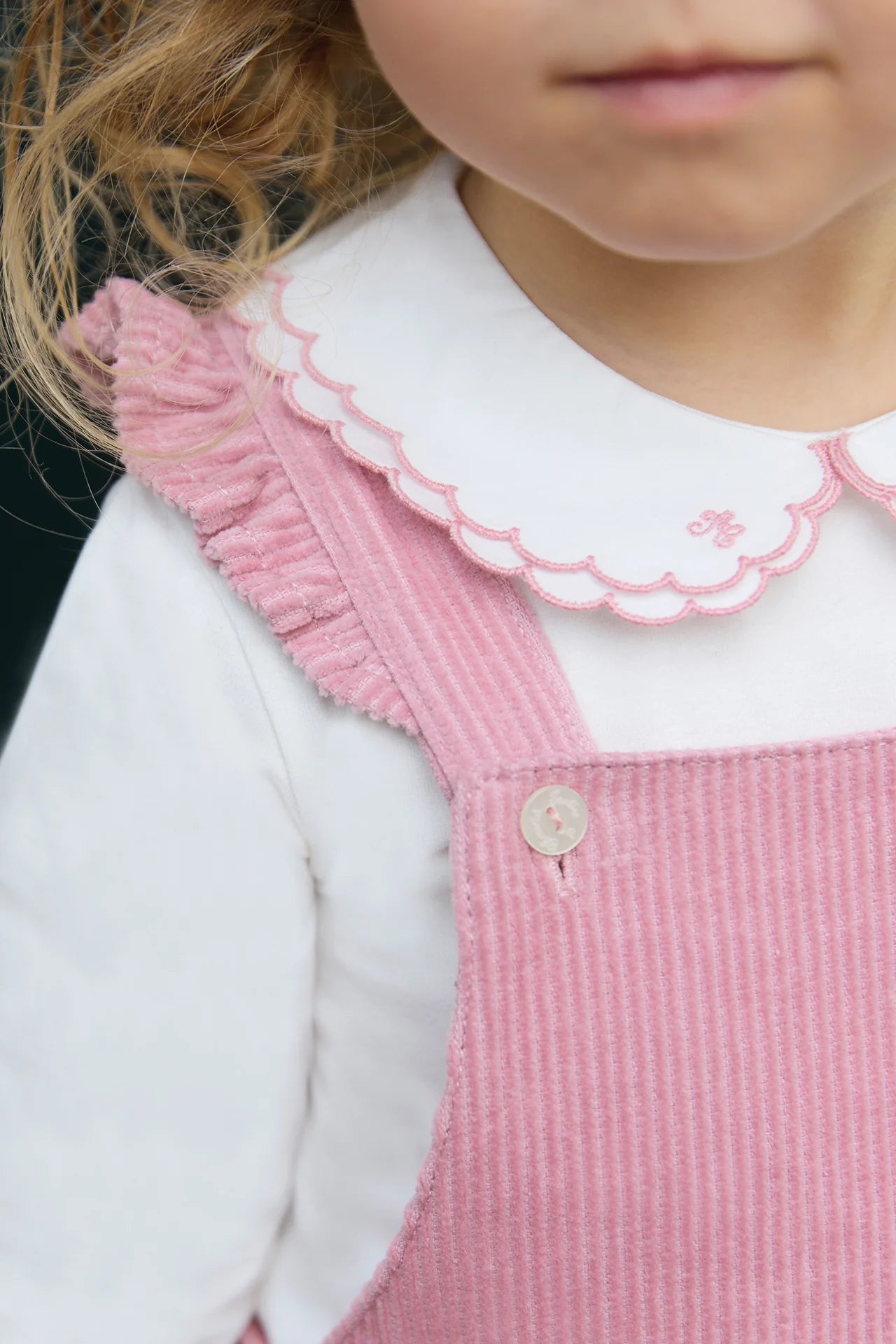 Close-up of a young child in Tartine et Chocolat’s Baby Girl’s Velvet Overalls in eglantine pink, featuring charming ruffle straps, worn over a white scallop-edged collared shirt.